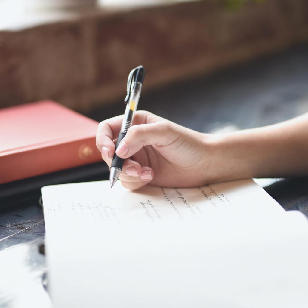 a hand holding a pen in an open book with a few words written down. a closed orange book nearby.